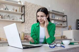 A young woman in glasses and green clothes is sitting at home in the kitchen at the table watching a movie, series, program from a laptop. She is crying, holding a napkin in his hands, wiping his tears. photo