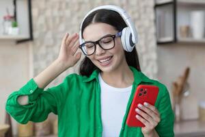 de cerca foto. contento joven hermosa hembra en verde ropa y lentes escuchando a música en blanco auriculares en el rojo teléfono a hogar en el cocina. ella cerrado su ojos y danzado a lo largo foto