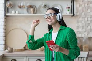 contento joven hermosa hembra en verde ropa y lentes escuchando a música en blanco auriculares en el rojo teléfono a hogar en el cocina. ella cerrado su ojos y danzado a lo largo foto