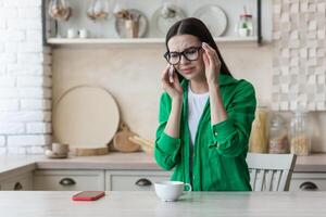 Depression, panic disorders. A young beautiful woman in glasses and a green shirt is crying at home, sitting at the kitchen table, wiping her tears with a napkin. He feels bad, feels fear and apathy photo