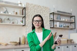 A young beautiful female nutritionist in glasses and a green shirt advises against eating chocolate. He holds a bar of chocolate and shows no with his hand. Chocolate and sweets are harmful to health photo