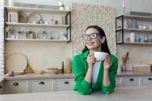 A young beautiful woman housewife in glasses and a green shirt rests from homework, takes a break, drinks coffee from a cup. Sitting in the kitchen at home, relaxed, thoughtful, smiling photo