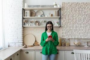 joven hermosa mujer en lentes y verde camisa en pie a hogar en el cocina mecanografía mensajes en el rojo teléfono, chateando con su amigos, utilizando un nuevo solicitud. foto