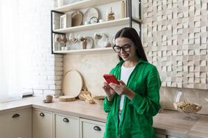 Young amorous teenage girl in glasses and green shirt texting with loved one in red phone at home. Embarrassed, she hides her phone, does not show it, smiles. photo