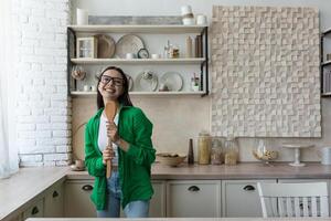 Young beautiful woman in glasses and green shirt dancing and singing at home in the kitchen. The housewife is preparing food, holding a spoon like a microphone. Resting from homework, happy, relaxed photo