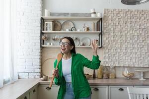 Young beautiful woman in glasses and green shirt dancing and singing at home in the kitchen. The housewife is preparing food, holding a spoon like a microphone. Resting from homework, happy, relaxed photo