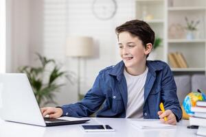 A young guy, a student at home, sits at a desk with a laptop, does homework and studies, smiles contentedly, records online courses, remote learning. photo