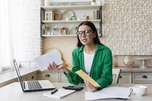 Mortgage loan. Upset and angry young woman holding a rent bill in her hands. Received a letter about the debt. Sittin g in the kitchen at home at the table with a laptop in glasses and a green jacket photo