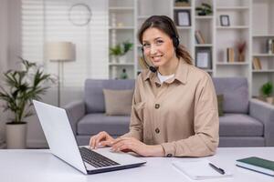 Young beautiful woman working from home remotely, businesswoman smiling and looking at camera, using headset phone and laptop for call, businesswoman inside home office. photo