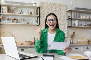 Happy young girl student in glasses and green shirt rejoices, received letter of invitation to university. received notification of admission. He sits at the table at home, opens an envelope. photo