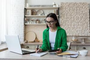 Home accounting.A young woman counts her income and expenses.checks the household budget. In glasses and a green shirt, sits at home with a laptop,a calculator and writes in a notebook,calculates. photo