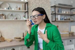 A young beautiful woman suffers from asthma, has seasonal allergies, uses an inhaler. At home in the kitchen, wearing glasses and a green shirt. photo