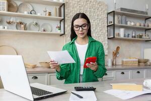 Work at home. A beautiful young businesswoman accountant works at home in the kitchen, sits at the table with a laptop and a calculator. She holds documents and a red phone in her hands. photo