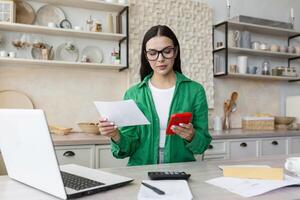 trabajo a hogar. un hermosa joven mujer de negocios contador trabajos a hogar en el cocina, se sienta a el mesa con un ordenador portátil y un calculadora. ella sostiene documentos y un rojo teléfono en su manos. foto