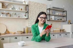 In love and happy young beautiful woman sends and receives love messages on the red phone from a loved one, from a boyfriend. Standing in the kitchen at home in glasses and a green shirt. photo