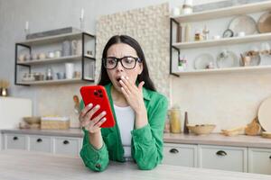 A young woman looks shocked and surprised at the phone, covered her mouth with her hand, saw a message from her ex-lover, learned about betrayal. Sitting at home in the kitchen and holding a red phone photo