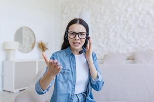 Webinar. Young beautiful business woman in glasses and headphones sits on the sofa at home and conducts a webinar, online training on camera, consults, tells, gestures with hands, smiles. photo
