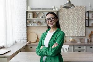 Portrait of a young beautiful brunette woman in glasses and a green shirt. Standing in the kitchen at home, looking at the camera, smiling, folded her arms over her chest. photo