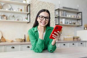 en línea compras. satisfecho hermosa joven mujer hace en línea compras, pedidos hogar entrega desde el rojo teléfono. él es sentado en el cocina a hogar en lentes y verde ropa, sonriente. foto