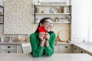 In love and happy young beautiful woman sends and receives love messages on the red phone from a loved one, from a boyfriend. Standing in the kitchen at home in glasses and a green shirt. photo