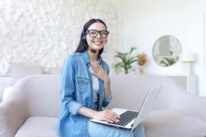 cuarentena. un joven hermosa mujer es sentado en un sofá con un ordenador portátil y vistiendo auriculares, felizmente hablando en línea con familia y amigos en un llamar, aislado a hogar en cuarentena. foto