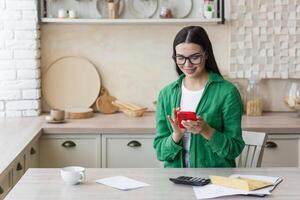 satisfecho joven hermosa mujer de negocios trabajando a hogar en cocina con rojo teléfono, documentos y calculadora. sentado a el mesa cerca el ventana en lentes y un verde camisa. foto