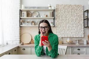 satisfecho hermosa joven mujer en lentes y un verde camisa a hogar en el cocina participación un rojo teléfono en su manos, contento en línea comprador, exitoso compras. foto