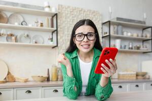nervioso joven mujer en lentes participación un rojo teléfono y mirando adelante a un importante llamada o mensaje. a hogar en el cocina, mirando a el cámara, preocupado. foto