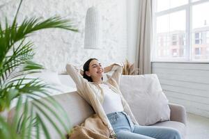 A young beautiful business woman is resting on the sofa after a successful working day. Rest break. She sits contentedly at home by the window with her eyes closed, hands behind her head, smiling. photo