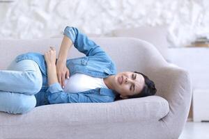 Menstrual pain. Young beautiful woman is lying on the sofa at home and holding her stomach. Feels pain on critical days. photo