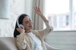 Beautiful young woman listening to music in white headphones. He sits at home on the sofa with his eyes closed, dances, sings, waves his hands. photo