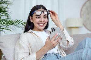 Happy young woman in glasses sitting on sofa at home, holding mobile phone, reading message, got good news. Satisfied and surprised, she lifted her glasses from her face with her hand. He smiles. photo