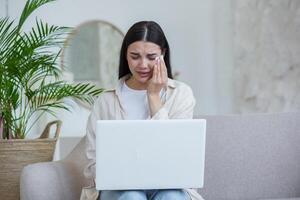 Quarantine. War. A beautiful young woman sits at home on the couch with a laptop, talks online via link with her family and friends, cries, misses them. Wipes tears with a napkin. photo