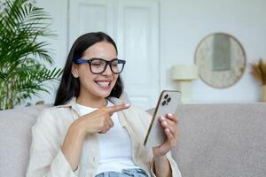 contento joven estudiante niña en lentes sentado en sofá a hogar, participación teléfono, chateando y chateando con amigos, sonriente. foto