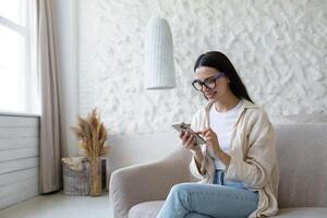 Blogging. Freelancer. Satisfied young beautiful woman in glasses sits at home on the sofa by the window and uses the phone, types, writes a post, blogs. photo