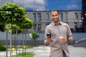 A person is holding a smartphone and a coffee cup, standing outdoors with trees and buildings in the background photo