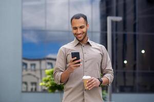 Businessman walking on break outside office building, male holding phone in hands, using app on smartphone to browse social network, holding cup of hot coffee drink in hand, smiling joyfully. photo