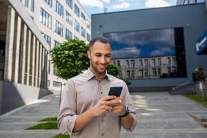 A young successful male businessman in a shirt walks outside an office building, holds a phone in his hands, uses an application on a smartphone, reads online pages, smiles happily photo