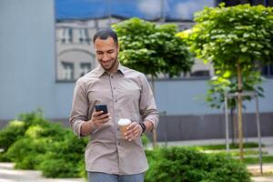 Businessman walking on break outside office building, male holding phone in hands, using app on smartphone to browse social network, holding cup of hot coffee drink in hand, smiling joyfully. photo
