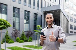 A man in shirt stands in front of a modern office building, holding a mobile phone and showing a thumbs-up gesture photo