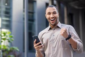 retrato de alegre africano americano alumno, hombre sonriente y mirando a cámara fuera de oficina edificio, participación teléfono won en línea ganar notificación, celebrando alto logro resultados. foto