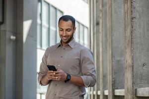 sonriente africano americano empresario en casual camisa utilizando un móvil teléfono fuera de un contemporáneo oficina estructura. foto