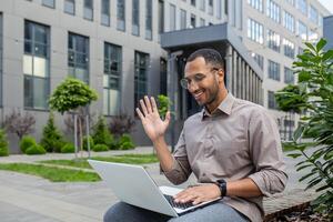 Businessman using laptop for call, man sitting on bench outside office building, waving to interlocutor, greeting gesture. photo