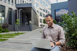 Smiling man with glasses using laptop outdoors, sitting by office building. Concept of remote work, technology, and happiness. photo