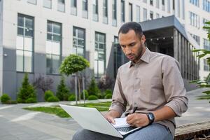 Focused young man using a laptop outdoors with a modern building backdrop, embodying mobile work and concentration. photo