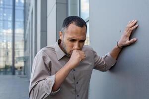 African American businessman appears to be sick, coughing while walking near a contemporary office structure. photo