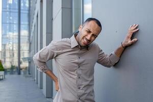 Man in casual shirt suffering from sudden lower back pain outside, grimacing in discomfort, hand on wall for support, expressing pain. photo
