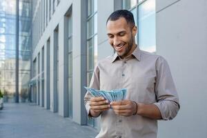 Smiling man counting money outdoors near modern office building, expressing financial success and prosperity photo