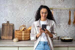 un joven hermosa mujer a hogar en cocina sonrisas contentamente, navega social redes, en línea Internet páginas, sostiene teléfono en r manos. Hispano con Rizado pelo , utilizando un aplicación en teléfono inteligente foto