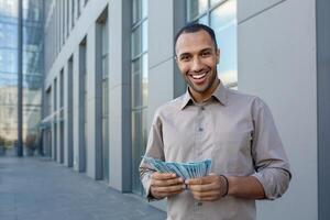 confidente africano americano masculino profesional en casual vestir participación dinero, expresando satisfacción y éxito fuera de un oficina edificio. foto